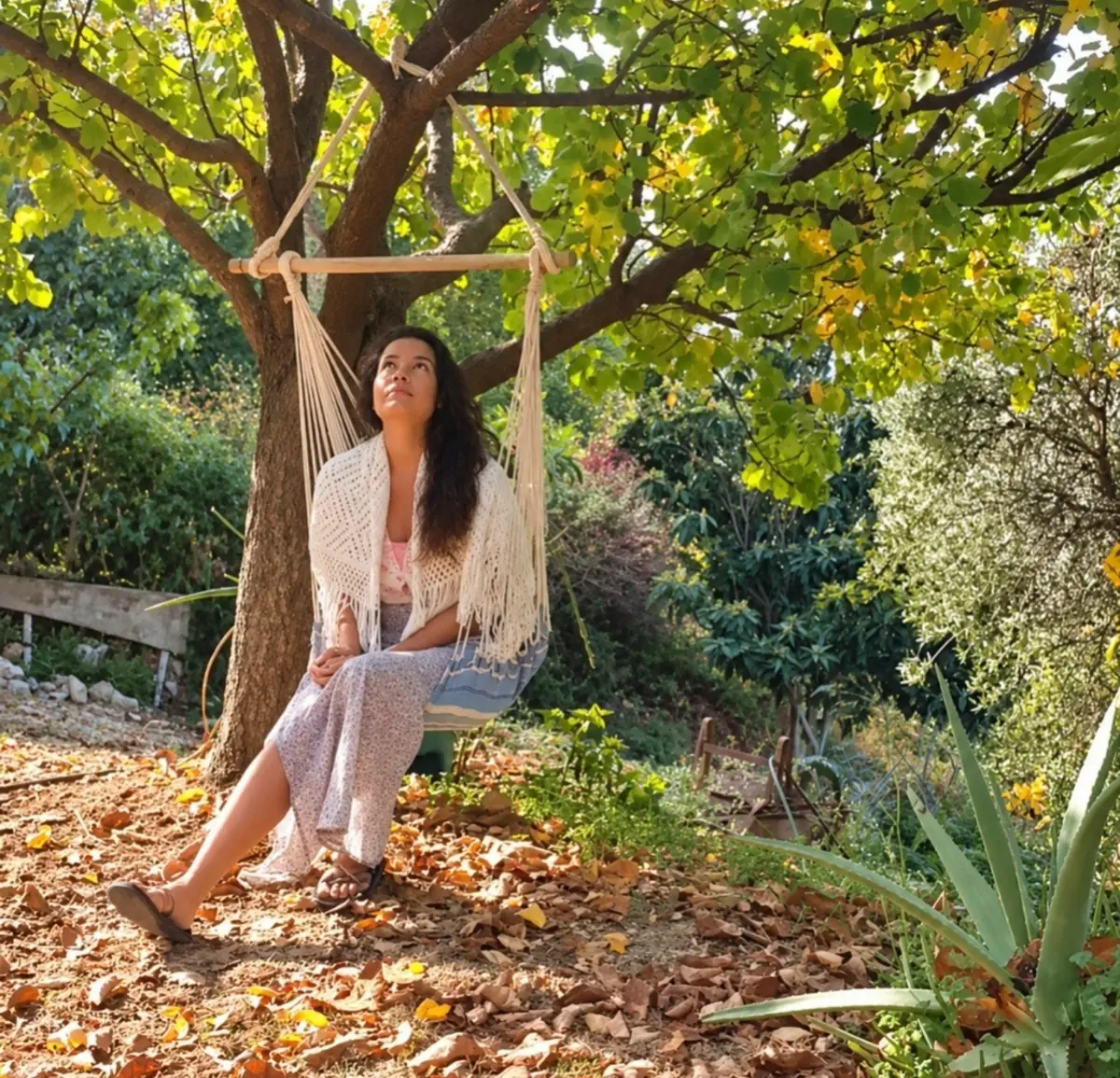 Dewi sitting in a hanging chair under a tree in autumn light, looking up, with a cream shawl around her shoulders.