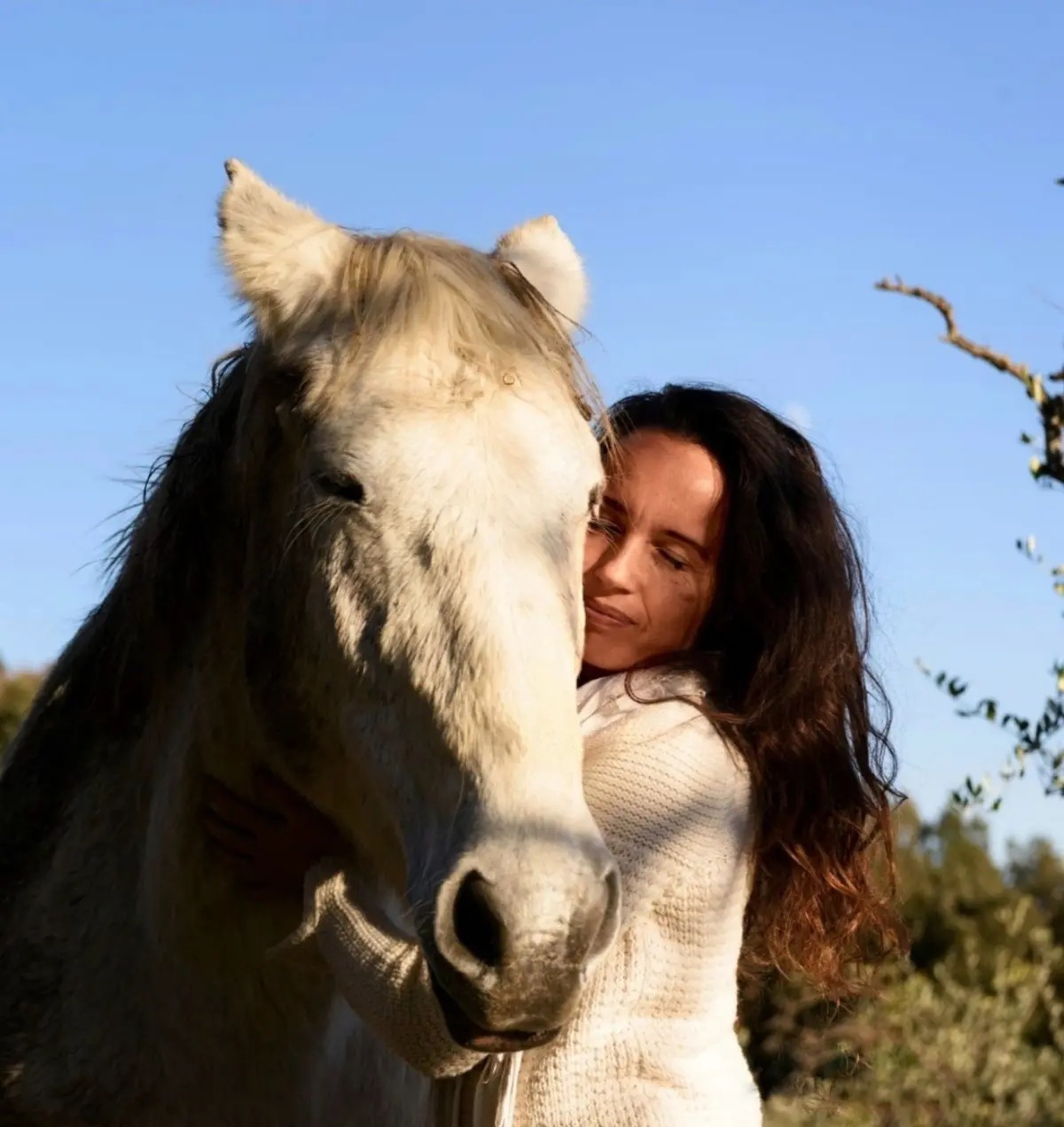 Dewi resting her cheek against the head of a white horse, eyes closed, under a clear blue sky.
