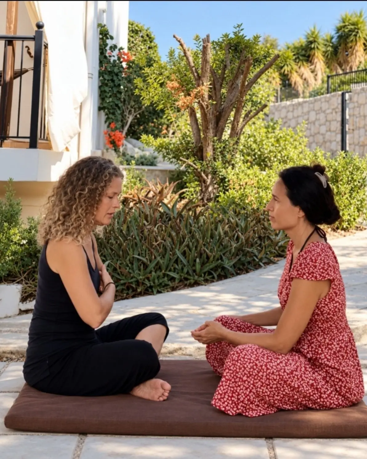 Two women sitting facing each other on a cushion in a sunlit garden, in quiet conversation.