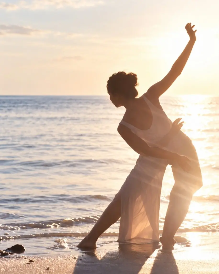 Dewi dancing at the water’s edge at sunset, one arm raised towards the light.