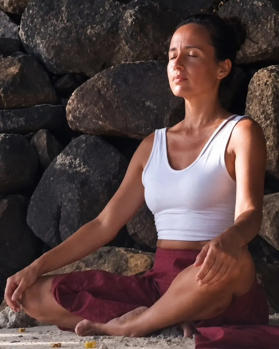 Dewi sitting cross-legged on pale sand against dark stones, eyes closed, in stillness.